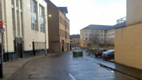 A general view of a street. There are green wheely bins on the road, with buildings on either side of the street. 
