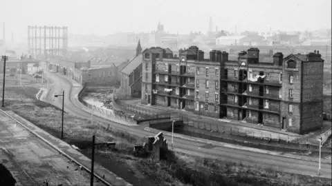 Manchester Libraries A black and white photograph of an industrial scene in Manchester with a four storey brick building and river flowing next to a road.