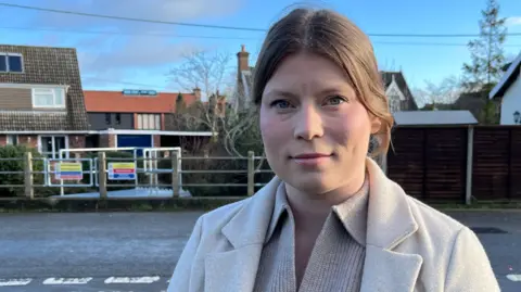 Shaun Whitmore/BBC A woman with brown hair tied back and wearing a beige jacket and a beige jumper. She is standing beside a road with houses in the background.