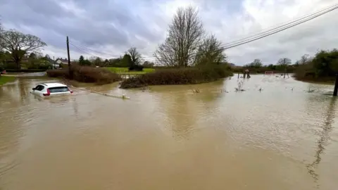 Martin Giles/BBC A white car is stuck in flood water. The water is above the tyres. A large area is covered by the water and houses can be seen in the background