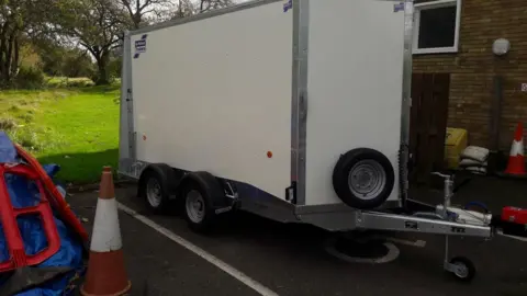 Hertfordshire Constabulary A grey trailer in a parking bay. The trailer is metal and not attached to a vehicle. To its right are parking cones and discarded road signs.