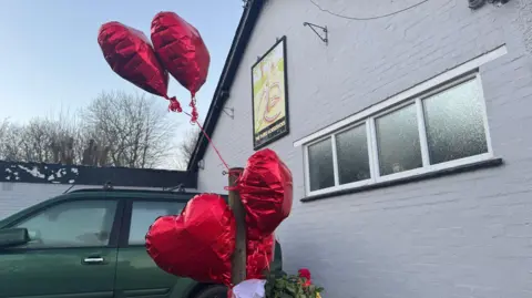 BBC/Jo Burn Burn empat balon berbentuk hati ditambatkan ke tiang gerbang di luar pub Three Horseshoes di Knockholt. Mobil hijau diparkir di belakang balon.