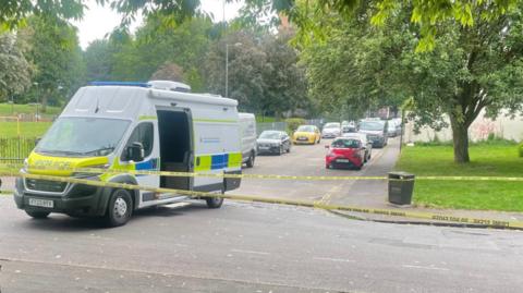 A police van behind yellow police tape, cordoning off a road next to a park.