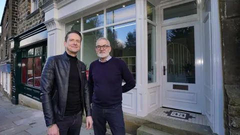Dan Merrick and Russell Trudgen stand on a stone-paved sidewalk in front of a building with a white-painted storefront. The building features large glass windows framed in white wood, and a white door with a rectangular glass panel. Below the door, there is a tiled entrance step with a decorative black-and-white mosaic pattern that includes the number “33”. The exterior walls of the building are made of dark stone, and adjacent to the white storefront is another shopfront painted in dark green.