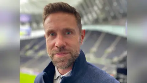 PA Media Matthew Upson, who has short brown hair and is starting at the camera with a slight smile. He has a blue quarter zip jumper on with a grey polo shirt underneath. He is standing in the Tottenham Hotspur Stadium with a large stand behind him, which is blurred in the background.