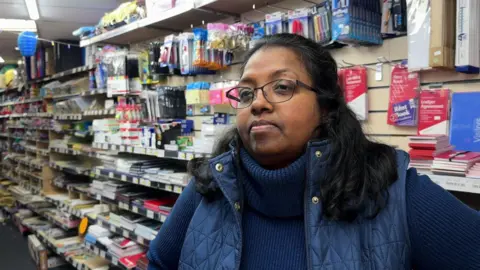 A woman wearing a dark blue gilet and turtle-neck jumper is standing inside a shop.