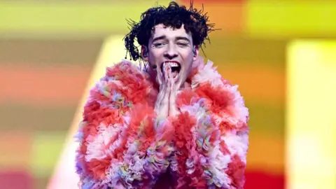 Singer Nemo celebrates after winning the Eurovision Song Contest. Nemo is wearing a pink and red ruffled coat