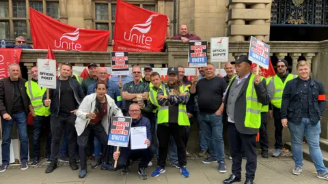 A large group of Unite union members are stood outside Sheffield Town Hall. They are wearing fluorescent tabards and carrying banners and placards.