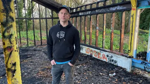 Steve Farmer pictured at the proposed site of his community cycle park at the former golf course at Broadwater Park, Farncombe. Steve is stood with his hands in his pockets and is wearing jeans, a black hoodie and a cap. 