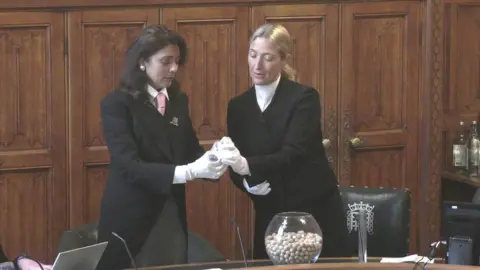 House of Commons A still showing Nusrat Ghani, the senior deputy speaker, being handed a ballot ball by a parliamentary clerk. They are standing in a wood panelled room in the Commons, between green leather chairs and a desk. On the desk sits a clear glass goldfish bowl, which is filled with small, white ballot balls. The two women wear formal black dress coats and white gloves. Ghani wears her black hair long and loose, whereas the clerk has her long blonde hair tied back in a ponytail. 