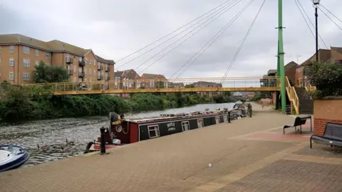 Canal path near Trenery Way in Northampton. There are canal boats in the water by the side of the path and a bridge with yellow railings across the water.