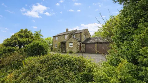 Paul Fosh Auctions The image shows a traditional stone-built house set within a green rural landscape. The property features stone walls and a slate roof. Attached to the right side of the house is a double garage with two brown doors. In the foreground, dense shrubs and leafy vegetation partially frame the view.