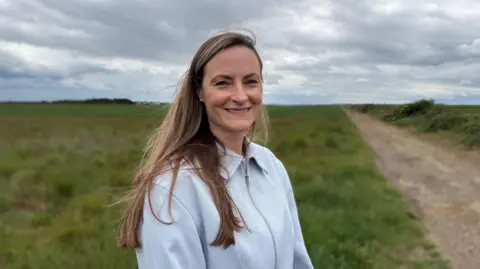 Steve Hubbard/BBC Natalie Ackroyd has long blonde hair and is wearing a light grey zip-up jacket. She is smiling and looking directly at the camera while standing in a field, next to a dirt track.