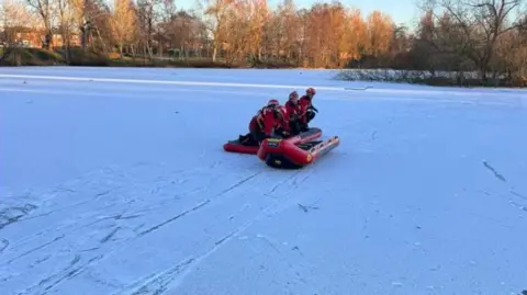 Carlisle East Fire Station View of an iced over pond, with a light covering of snow. Three members of Cumbria fire service wearing red high-viz jackets and helmets are on a sledge with an inflatable next to them. In the background trees are catching the winter sunlight.