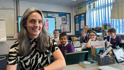 Andrew Turner/BBC Hannah Barrett has long silvery grey hair, and she is smiling. She is at a desk with children behind her. She is wearing a black-and-white patterned dress. Behind the children are blue vertical blinds at the window, and children's work is pinned to the walls. There are books on the desk in the foreground.