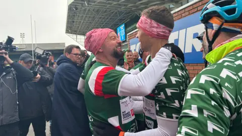 Kate Bradbrook/ BBC Two men in a green, white and red rugby tops embrace at a rugby ground as photographers take photos. 