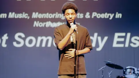 Project M A young person - likely a teenager or young man - performing into a microphone, wearing a dark T-shirt and trousers. He has one hand on a microphone and one on his stomach. Words are printed on a wall or screen behind him. A drum kit is to the right. 