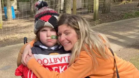 Bev Price A boy with long hair is wrapped in a wooly hat, football blanket and navy hoodie. He also has a medical tube up his nose and is sat in a wheelchair. A women in an orange jumper with long blonde hair is hugging him.