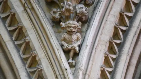 Getty Images Stone carving of the Lincoln imp with exaggerated facial features and pointed ears, seated with crossed legs and clasped hands, positioned within an ornate arch of the cathedral. 