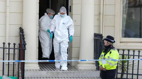 Eddie Mitchell Two officers leave the building wearing outfits associated with forensics officers 