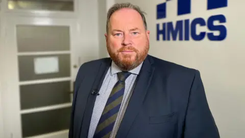 His Majesty's Inspectorate of Constabulary in Scotland Craig Naylor staring straight at the camera while standing in a room with a blue HMICS logo in the background 