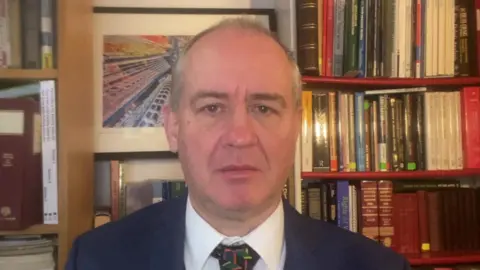 A man with very short white hair looks straight into the camera. He is wearing a Navy blue suit and white shirt. He is sitting in front of bookshelves full of books.