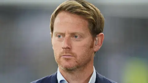 Michael Stewart has ginger hair and grown out stubble. He has a neutral expression and looks off to one side 