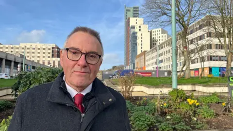 Tudor Evans, leader of Plymouth City Council, wearing a white shirt and red tie and a grey zipped coat, standing in Armada Way, Plymouth