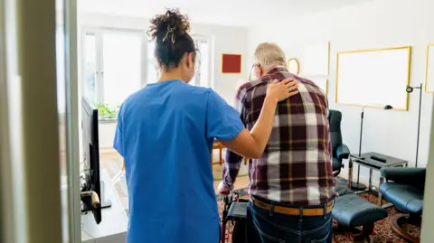 Getty Images A homecare worker is has a hand resting on an elderly man's back. He is using a zimmer frame to walk.  The worker is wearing blue overalls and the man is wearing a maroon and yellow checked shirt and jeans. They are in a living room. There's a TV, some tables and a chair in the background. 