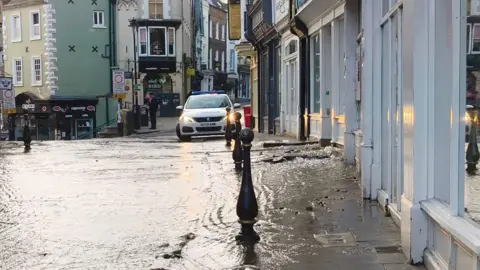 Claire Donlan Water flowing down Saddler Street in Durham. There are shop fronts on the right side of the narrow streets with restaurants and a police car in the distance.