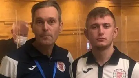 Steve and Byron Down standing next to each other in front of a wooden wall. Steve is wearing a navy tracksuit jacket and Byron is wearing a white polo shirt.