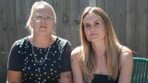 Shaun Whitmore/BBC Terri Bird, a blonde woman with glasses wearing a black patterned top, sits beside Layla Eales, who has long blonde hair and is wearing a black strappy vest top