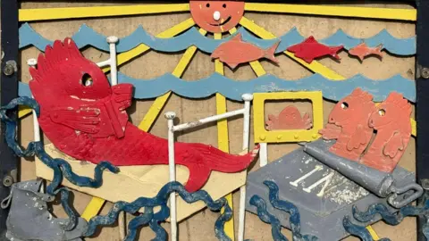 Carl Quartermain A design of a red fish lying on a deck chair next to a tin of sardines. The sea is behind them with the sun prominently in the middle.