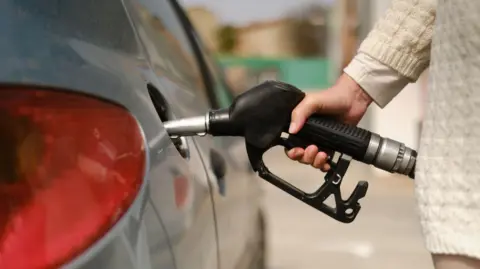 A woman's arm and hand holding a black petrol pump and using it to fill her silver car