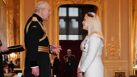 PA Media The King, dressed in a dark military uniform with gold detailing, stands facing Katherine Goodwin, who is wearing a light grey dress and a small black fascinator. They appear to be conversing during an investiture ceremony in an ornate gold-trimmed room, with a uniformed officer standing in the background.