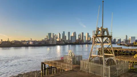 Historic England Archive A crane-like tower with gaps in stands inside a metal fence, in front of it is a smaller wheel shaped object inside a smaller metal fence. There is blue water with the view of a London cityscape in the distance, the sky is mainly blue with a peach strip low in the sky.