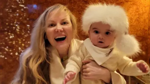 Lucy Turner Lucy is laughing at the camera while holding baby Sandy. She has long blonde hair and a white jumper on. Sandy is wearing a white jumper and has a white fluffy Christmas hat on that has a pompom.