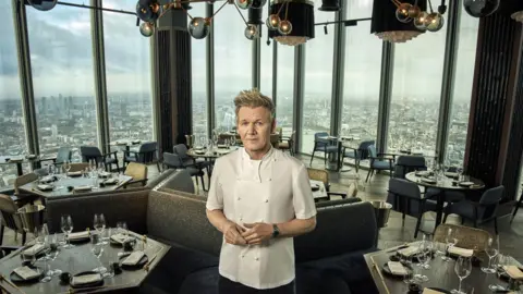 PA Media Gordon Ramsay stands in the middle of a high-rise restaurant with large windows showing a panoramic view of London. The restaurant has dark wooden tables set with wine glasses, plates, and napkins, surrounded by plush chairs and booths. The lighting fixtures above have a modern, gold-toned design.