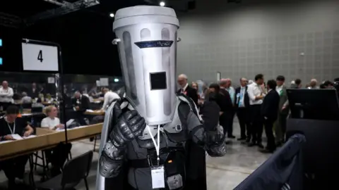 Getty Images A person in a black costume with a bin for a helmet posing for photos at an election count 