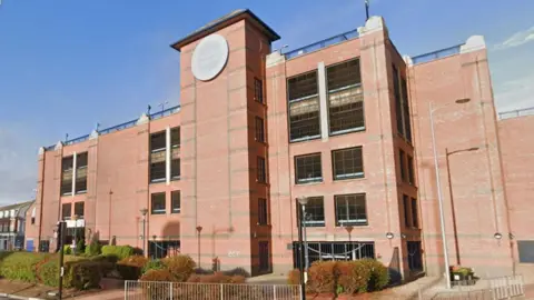 A photo of the exterior of Freshney Place shopping centre. The image shows a large, multi-story red brick building with a tall central tower. It has multiple rectangular windows, some covered with black grilles. A circular sign is on the tower reading Freshney Place. The foreground features bushes, lamp posts, railings and a paved walkway. The sky is clear blue.