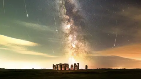 Josh Dury Stonehenge as seen from afar, with the base of the stones lit up slightly. Above the monument, you can see a large vertical cloud shape of white and purple, which is the Milky Way galaxy. Either side of the galaxy, thousands of stars can be seen, and meteors can be seen streaking towards the ground.