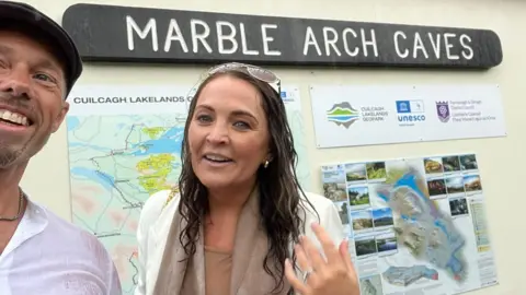 Alistair Hay Alistair Hay and Kim Mahon Hay standing in front of a sign for Marble Arch Caves. Alistair is wearing a flat cap, silver necklace and white shirt. Kim has long brown hair wearing sunglasses on top of her head, and a cream blazer. Both are soaked.