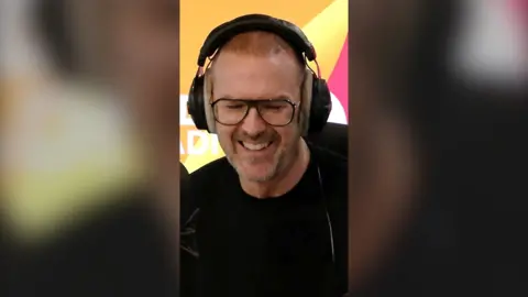 Paddy McGuinness wears headphones and black rimmed glasses, looking down, smiling