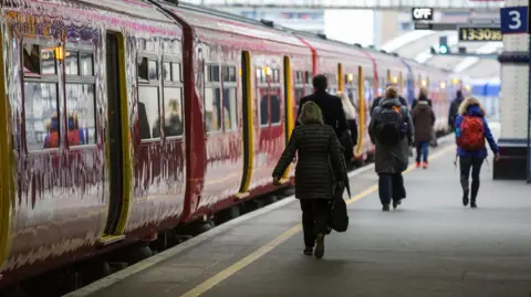 A red South Western Railway train at London Waterloo, Platform 3, with passengers walking alongside the train. A clock reads 13:30.