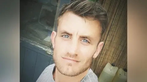 Paul Stewart Miller is taking a selfie and looking directly at the camera. He has short brown hair and facial hair, and is wearing a grey-striped shirt. There is some sort of filter on the photo that has brightened the colours and softened the edges.