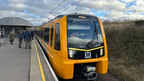 A yellow and black new style metro train at an outdoor station, with passengers waiting on the platform. Around building sits on the central platform between the two rail lines.