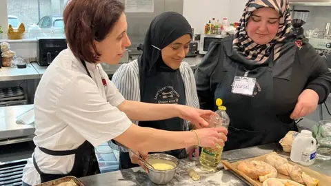 Denisa Theodorescu stands in a kitchen in chef's whites with a black apron over the top. She has thick, wavy dark hair and is holding a bottle of oil as a woman in a black headscarf holds a metal bowl next to some bread on a tray. They are watched by another woman in a black long-sleeved top and black and beige striped headscarf