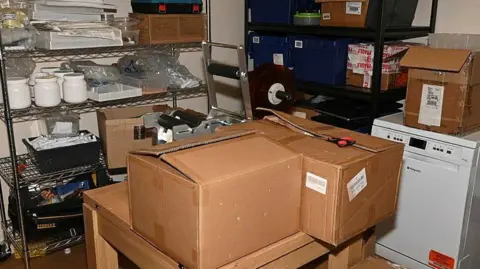West Yorkshire Police A cluttered storage and packing room containing cardboard boxes, shelving units and warehouse equipment. Several sealed boxes sit on a wooden table in the foreground alongside packing tools, including a tape dispenser and cutting tool. Metal shelving around the room holds containers, supplies, packaging materials and plastic storage bins, while additional boxes are stacked on shelves and the floor. A white appliance and various storage items are visible in the background, giving the space the appearance of a stockroom or logistics area.