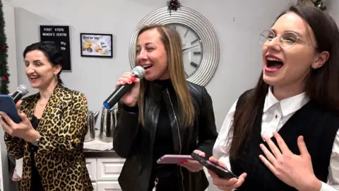 Svitlana, far left, is wearing a leopard-print jacket and singing into a microphone. She has dark hair in a pony tail. Her friends are beside her - one is a woman with long light-brown hair singing into a microphone and wearing a black jacket and black top. The other closest to the camera has dark brown hair and round glasses. She is wearing pink lipstick and has a white blouse on, over which is a black tank top.