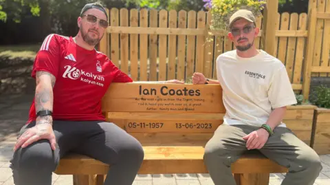 Two men sat on a wooden bench dedicated to Ian Coates. It reads "Ian Coates. As the mist rolls in from the Trent, fishing rod in hand you watch over us. A great father, family man and friend. 05-11-1957 13-06-2023"